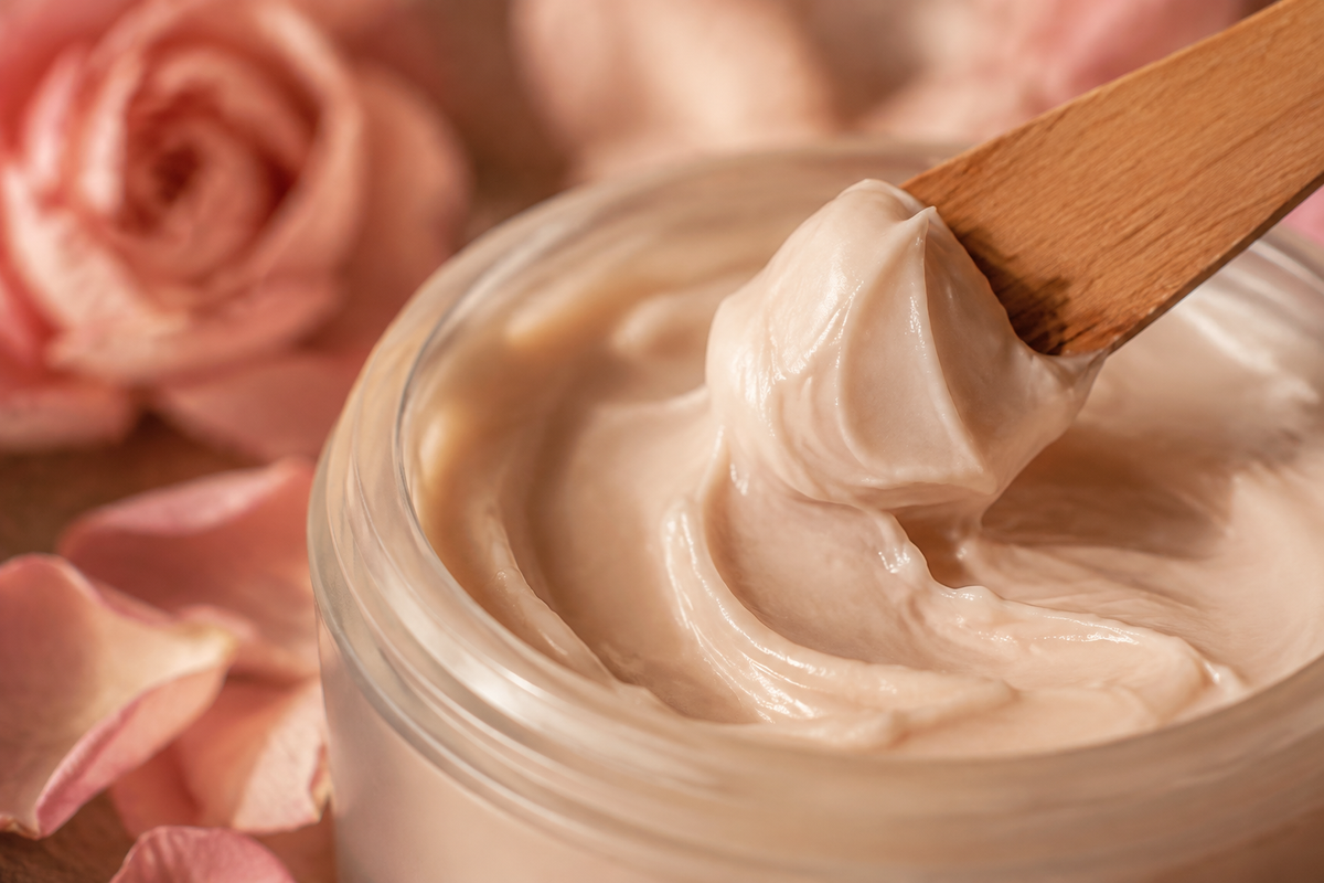 A close-up of creamy lotion texture with a wooden spatula and rose petals